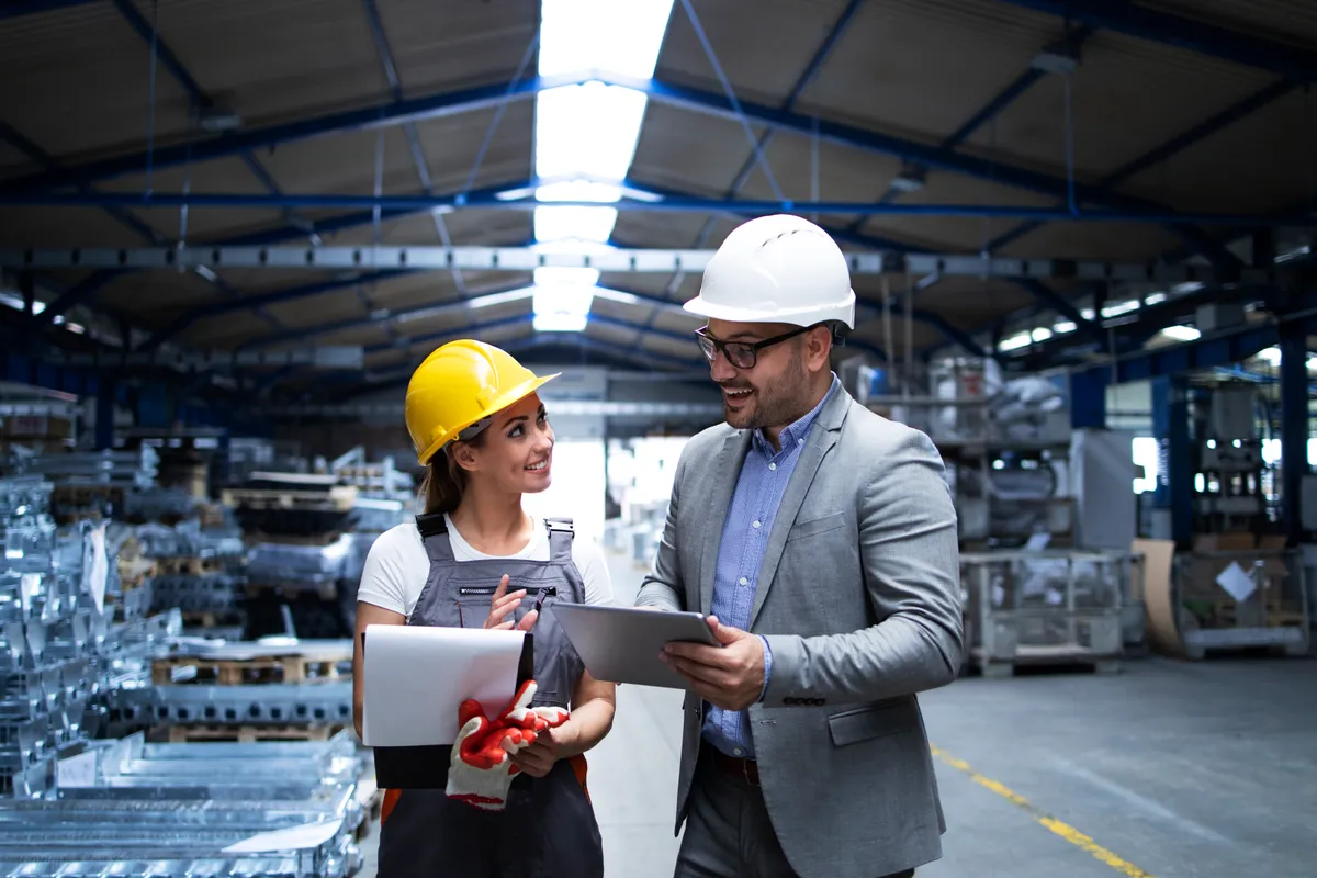 Two colleagues wearing safety helmets discussing documents and a tablet on a factory floor.